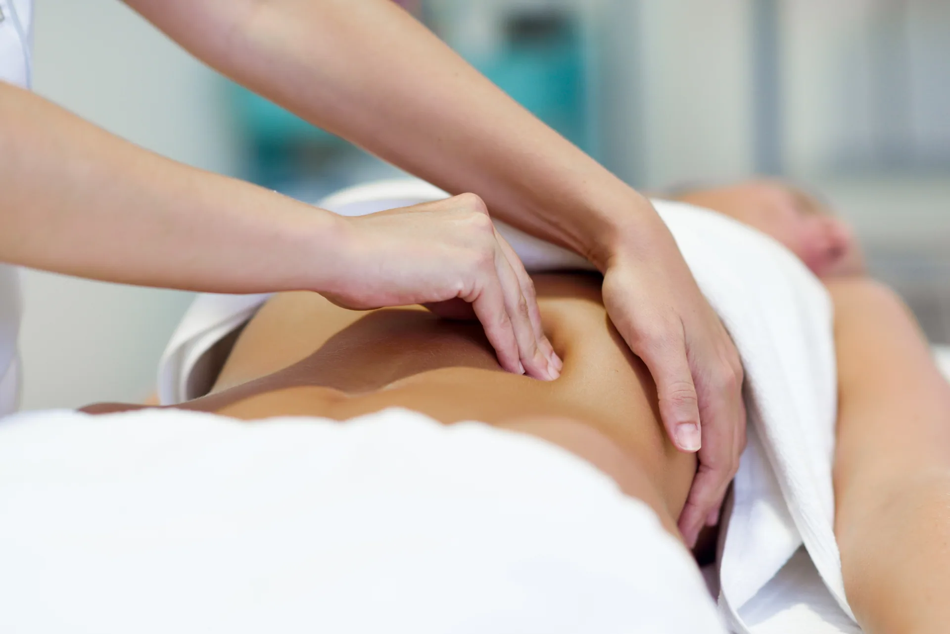 Therapist's hands performing an abdominal massage on a client lying on a treatment table draped in white towels.