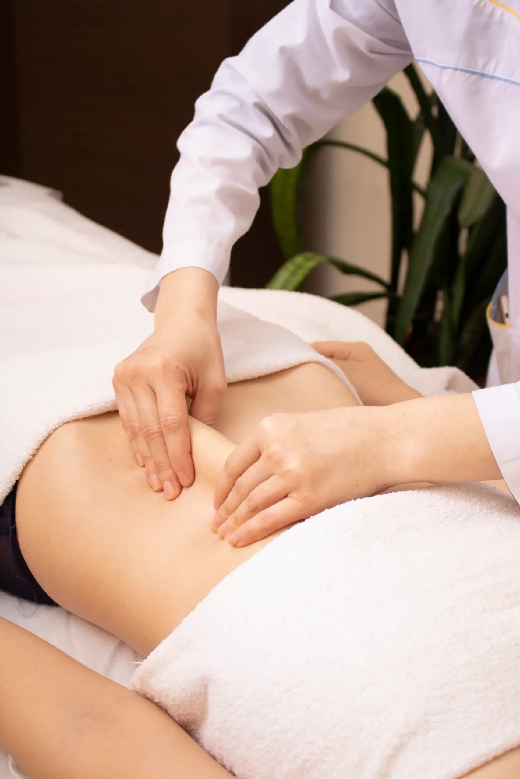 Therapist in a white coat presses fingers into a client's abdomen during a massage, client draped in white towels.