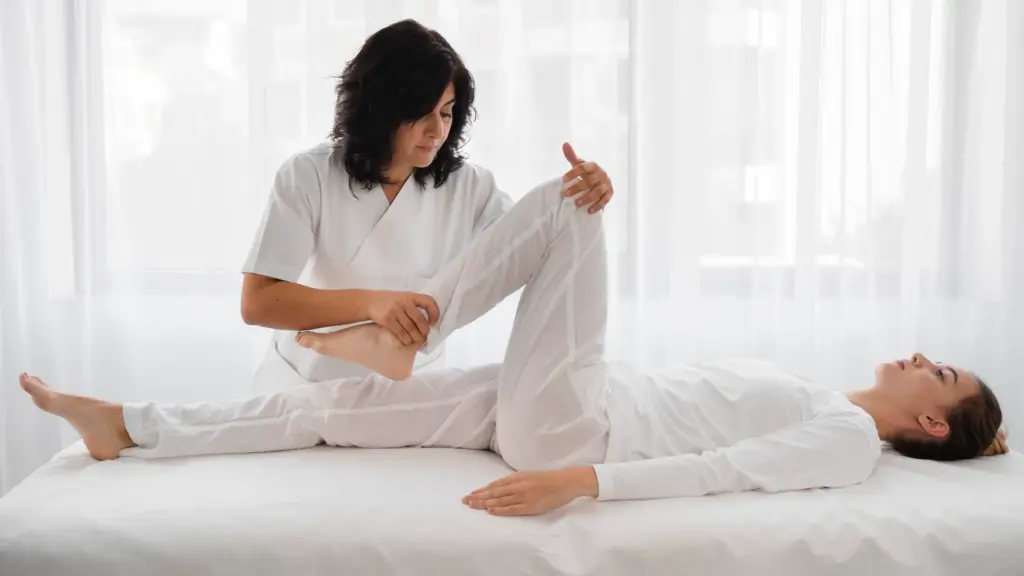 Massage therapist guides a client's bent leg in an assisted stretch while the client lies on a treatment table.