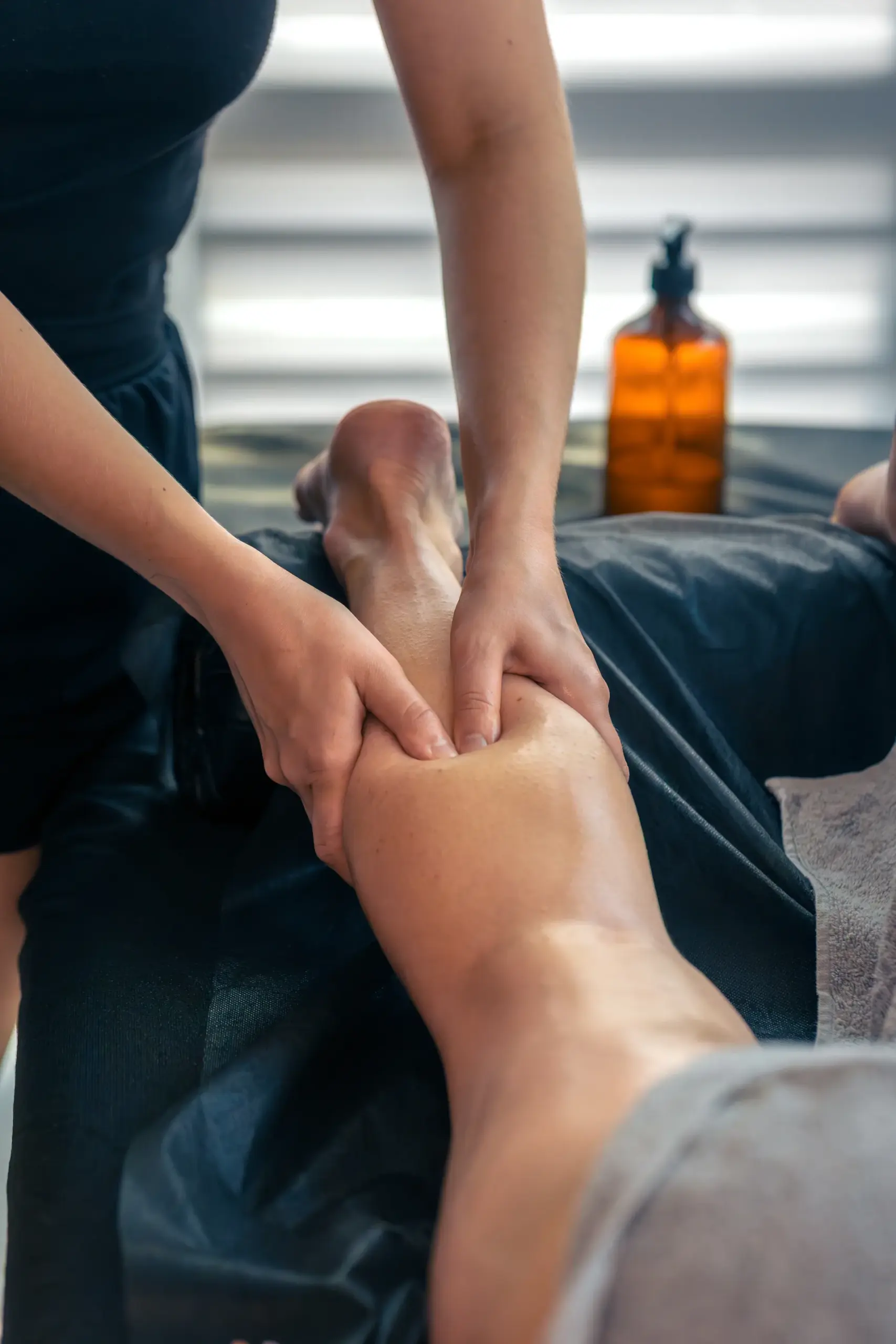 Massage therapist using both hands to work on a client's calf muscle on a massage table, amber oil bottle in background