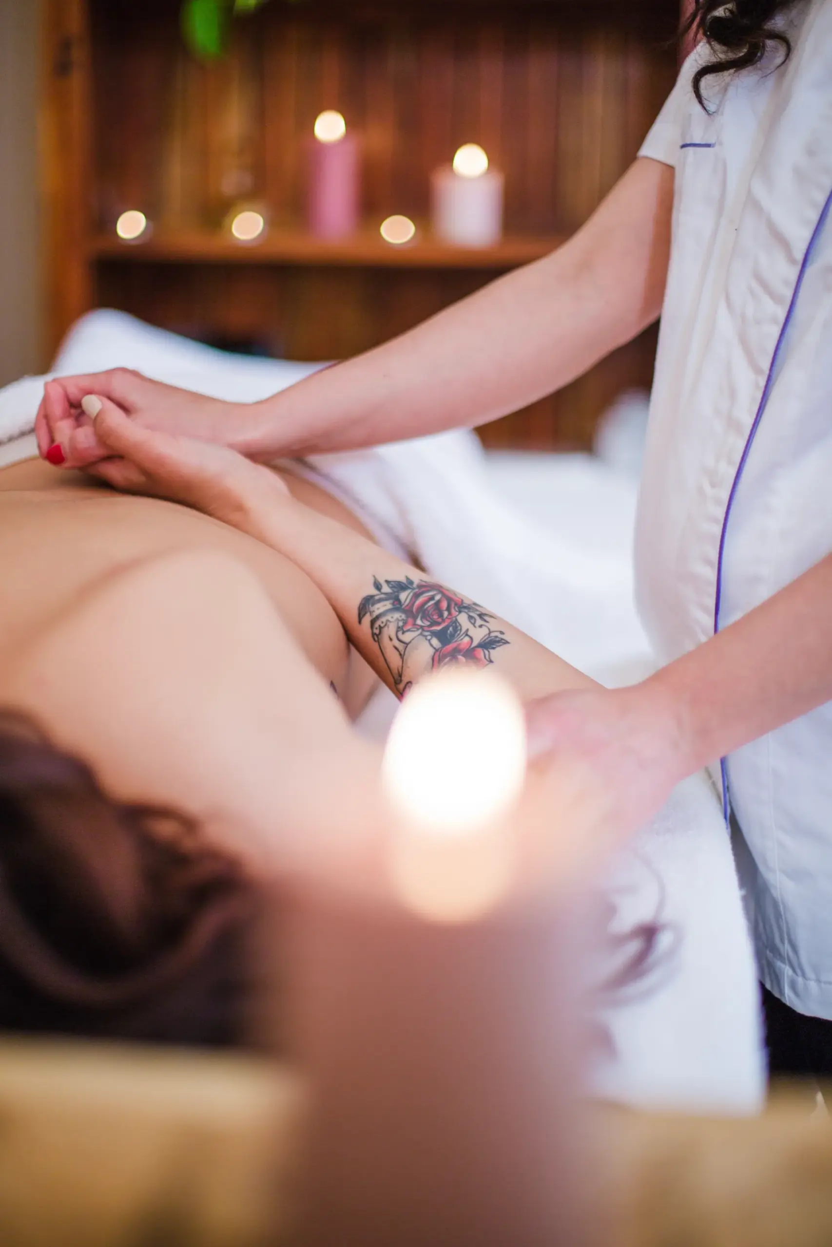 Massage therapist in a white tunic massaging a client's arm on a spa table with lit candles in the background.