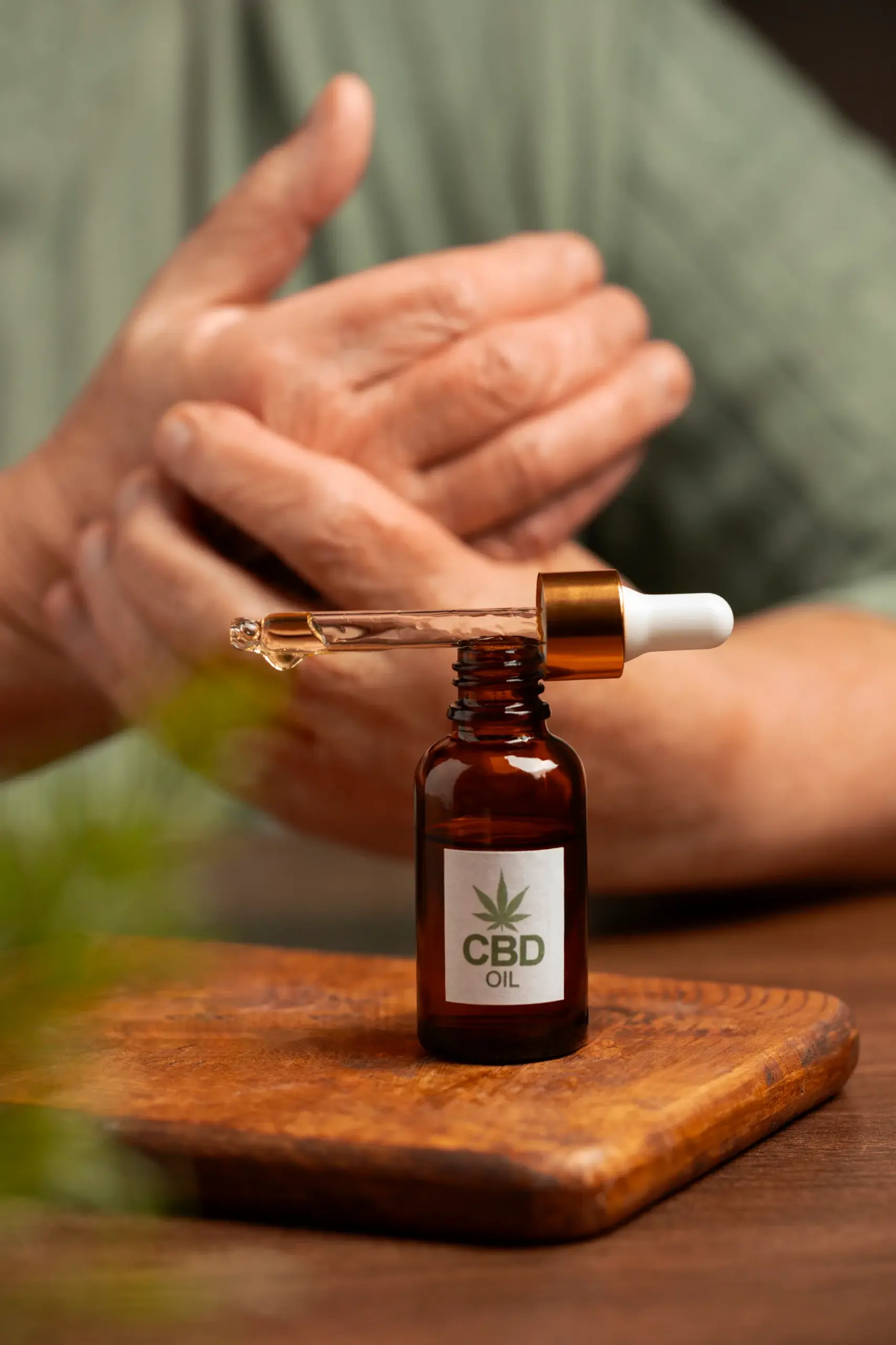 Amber CBD oil bottle with dropper on a wooden board, a person rubbing their hands in the background.