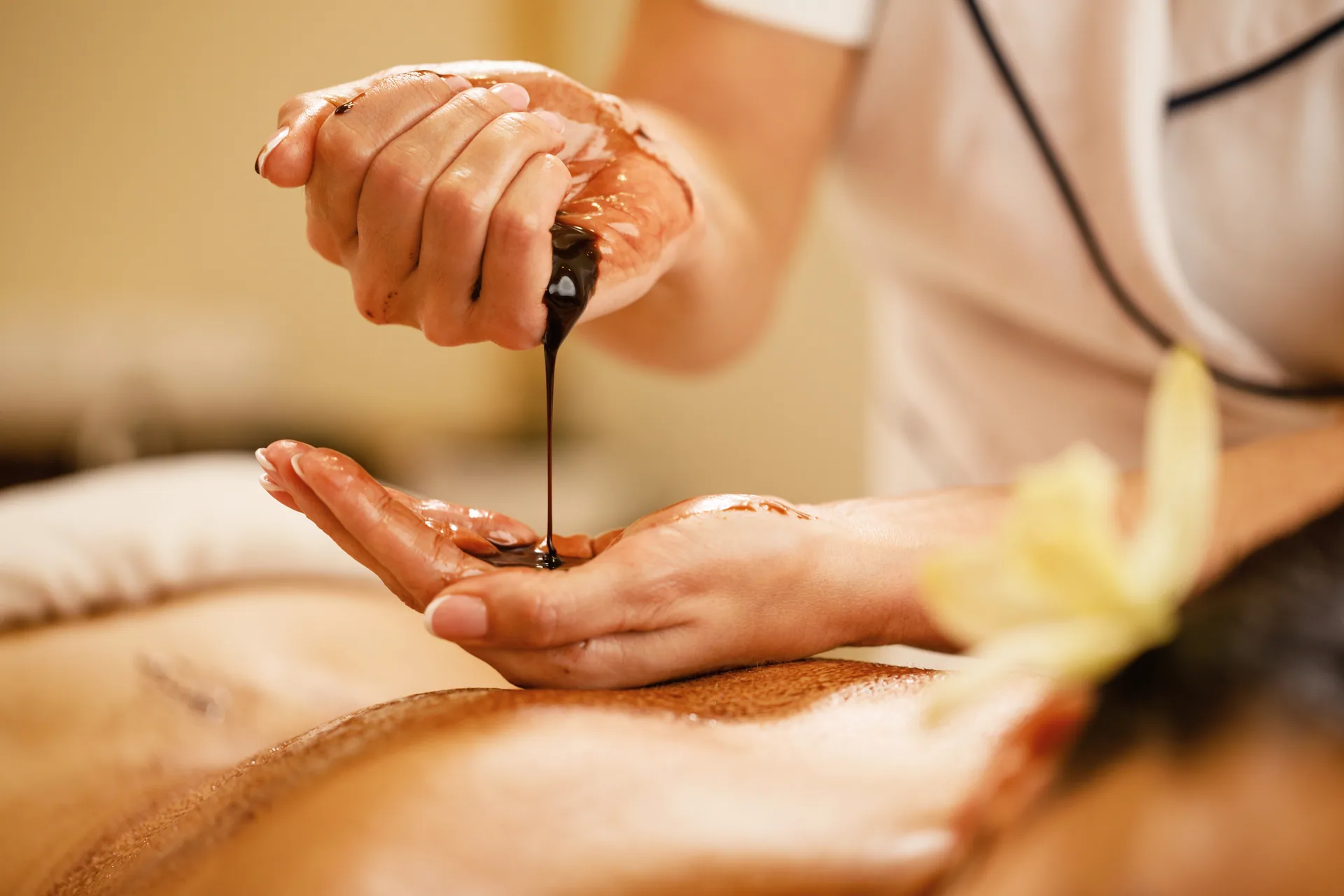 Massage therapist pouring warm chocolate oil from one hand into the other above a client on a spa table.