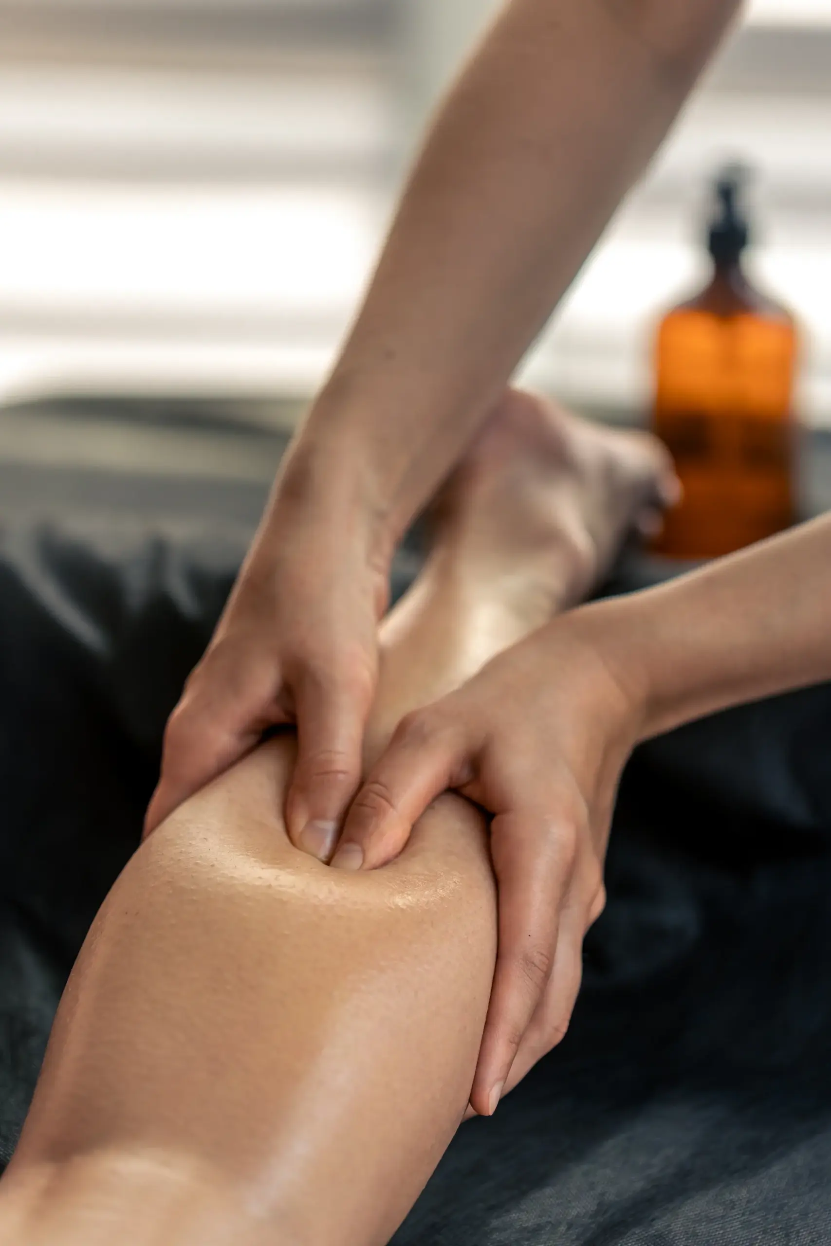 Therapist's hands pressing into a client's oiled calf during a leg massage, amber oil bottle in the background