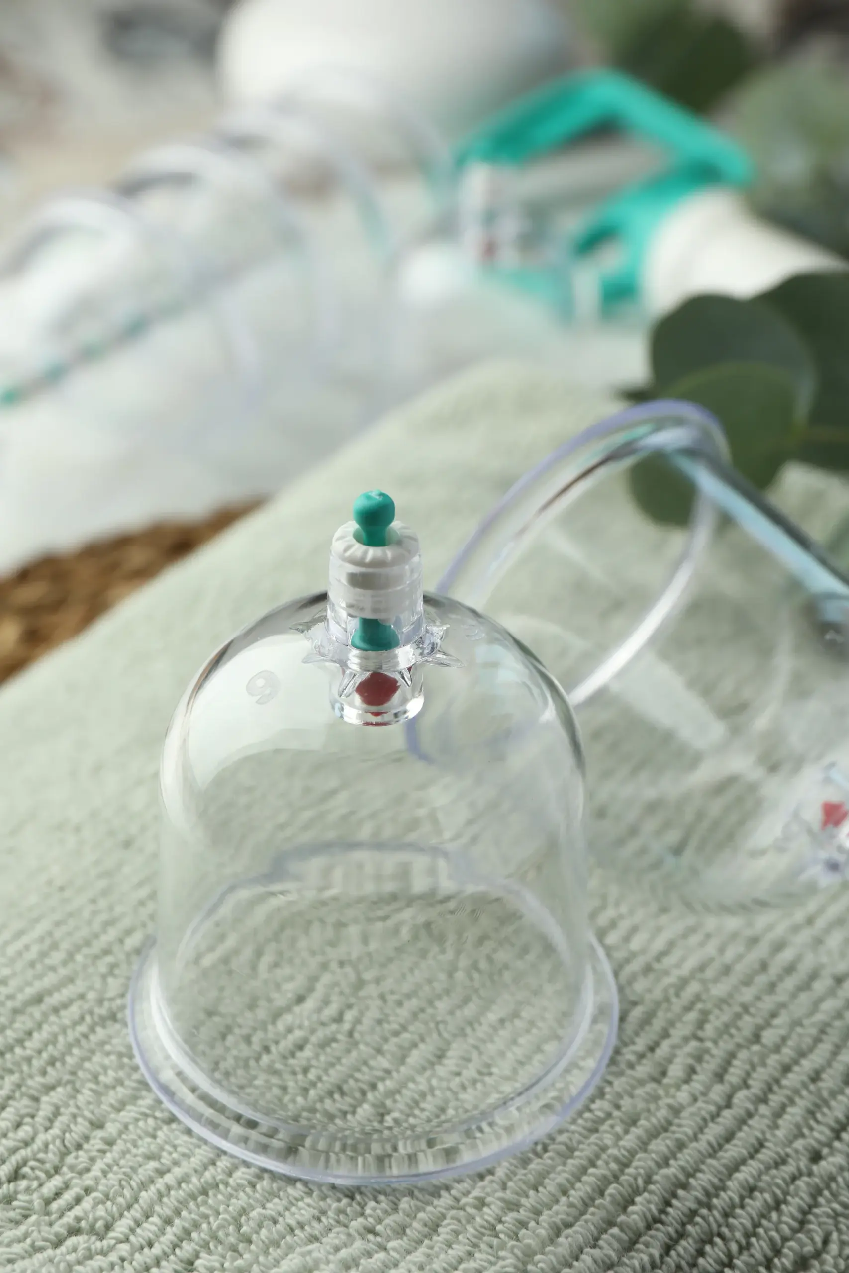 Close-up of a clear plastic cupping therapy cup with a valve top resting on a folded green towel, more cups blurred behind.