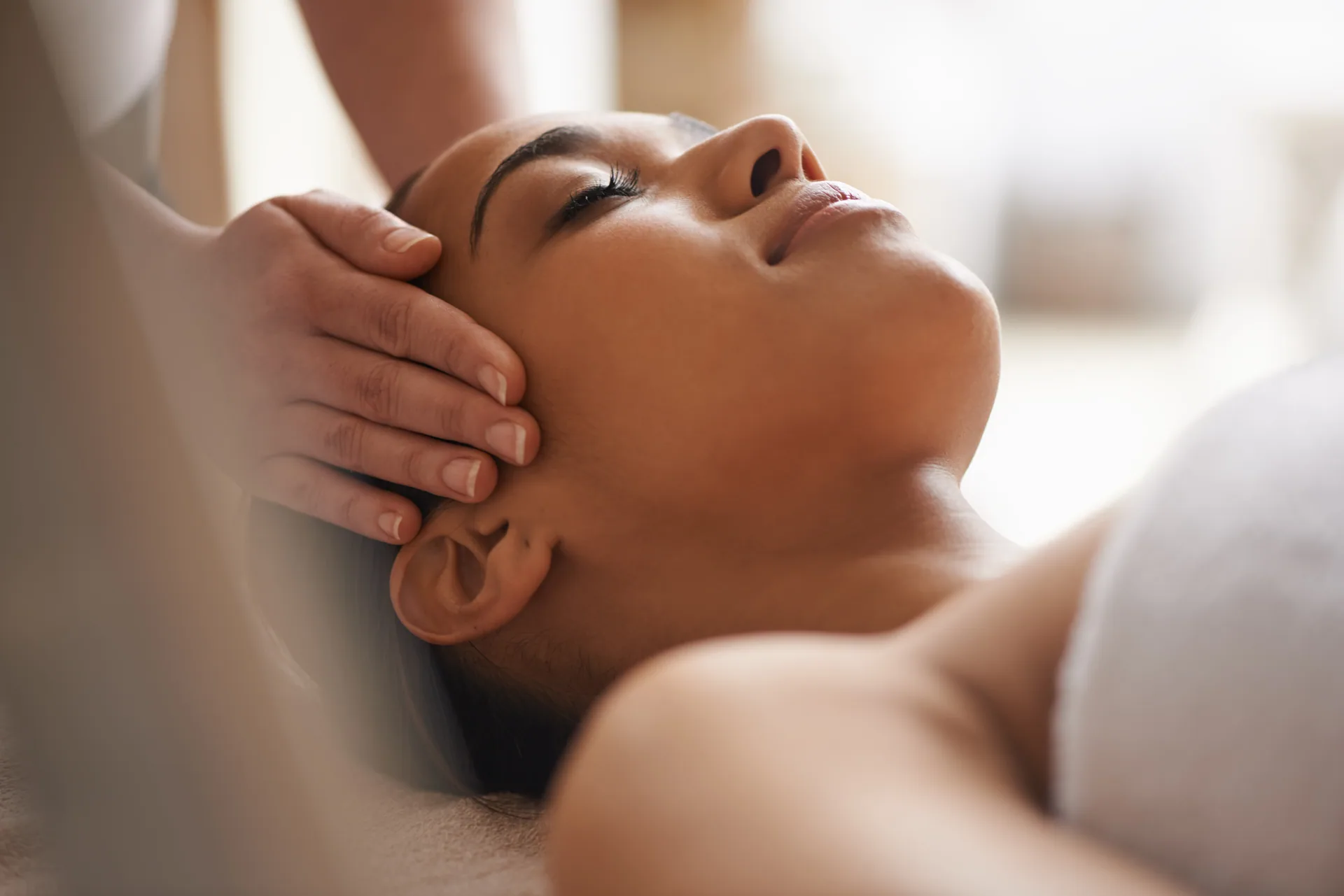 Close-up of a woman lying with eyes closed while a therapist's hands massage her temples and scalp.