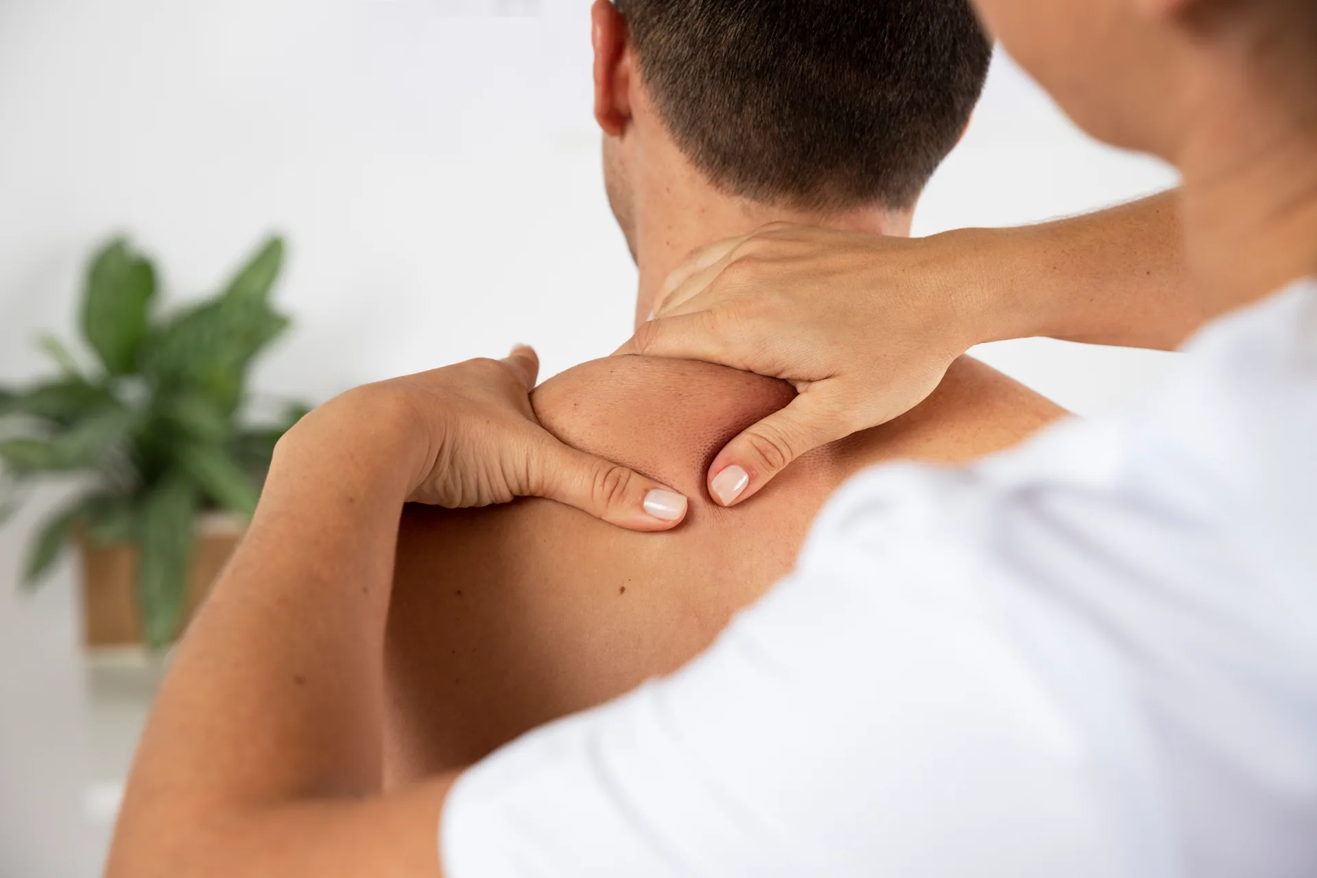 Massage therapist pressing both thumbs into a man's upper shoulder muscles during a treatment session.