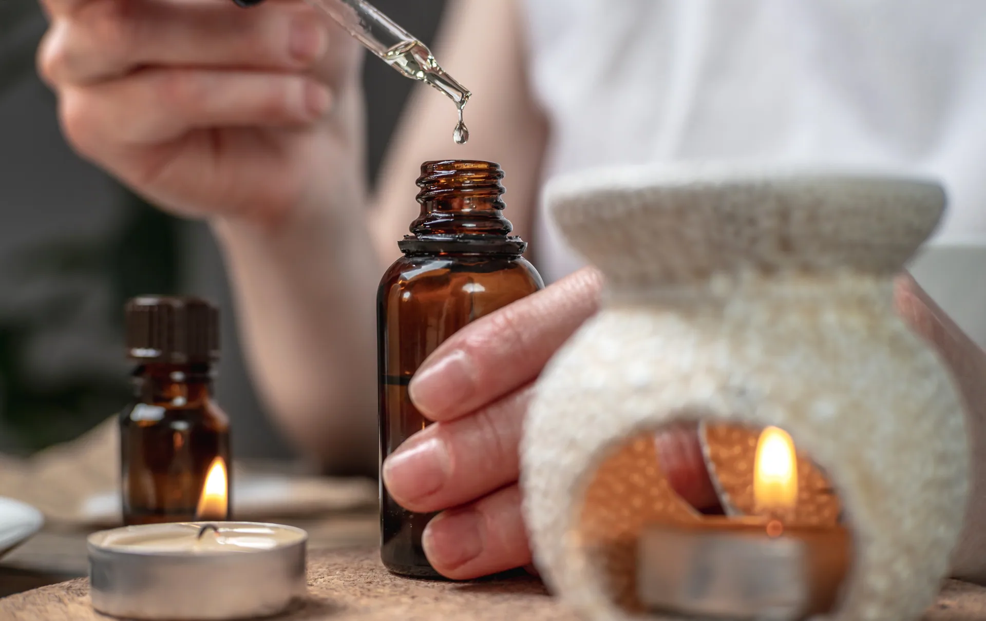 Hand releasing a drop of essential oil from a glass dropper into an amber bottle beside a lit candle and diffuser.
