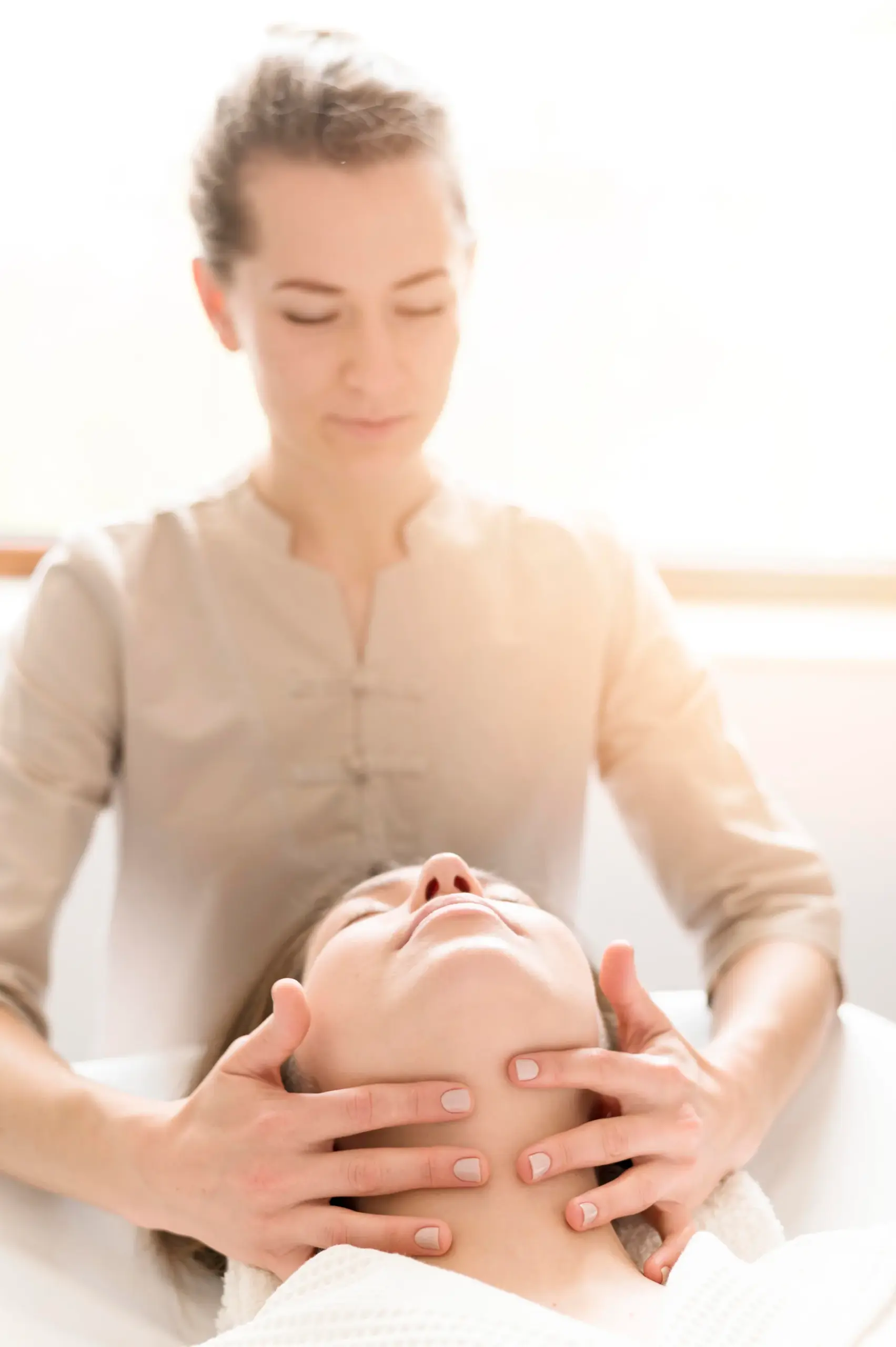 Therapist in a beige tunic massages the face and jaw of a client lying on a treatment table.