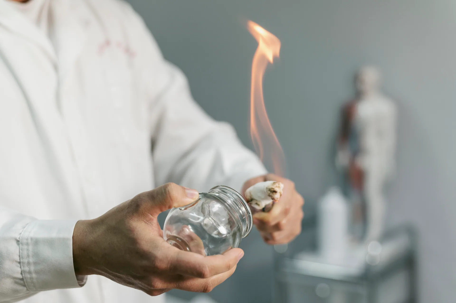 Practitioner in a white coat holds a flaming cotton swab beside a glass cup to prepare for fire cupping therapy.