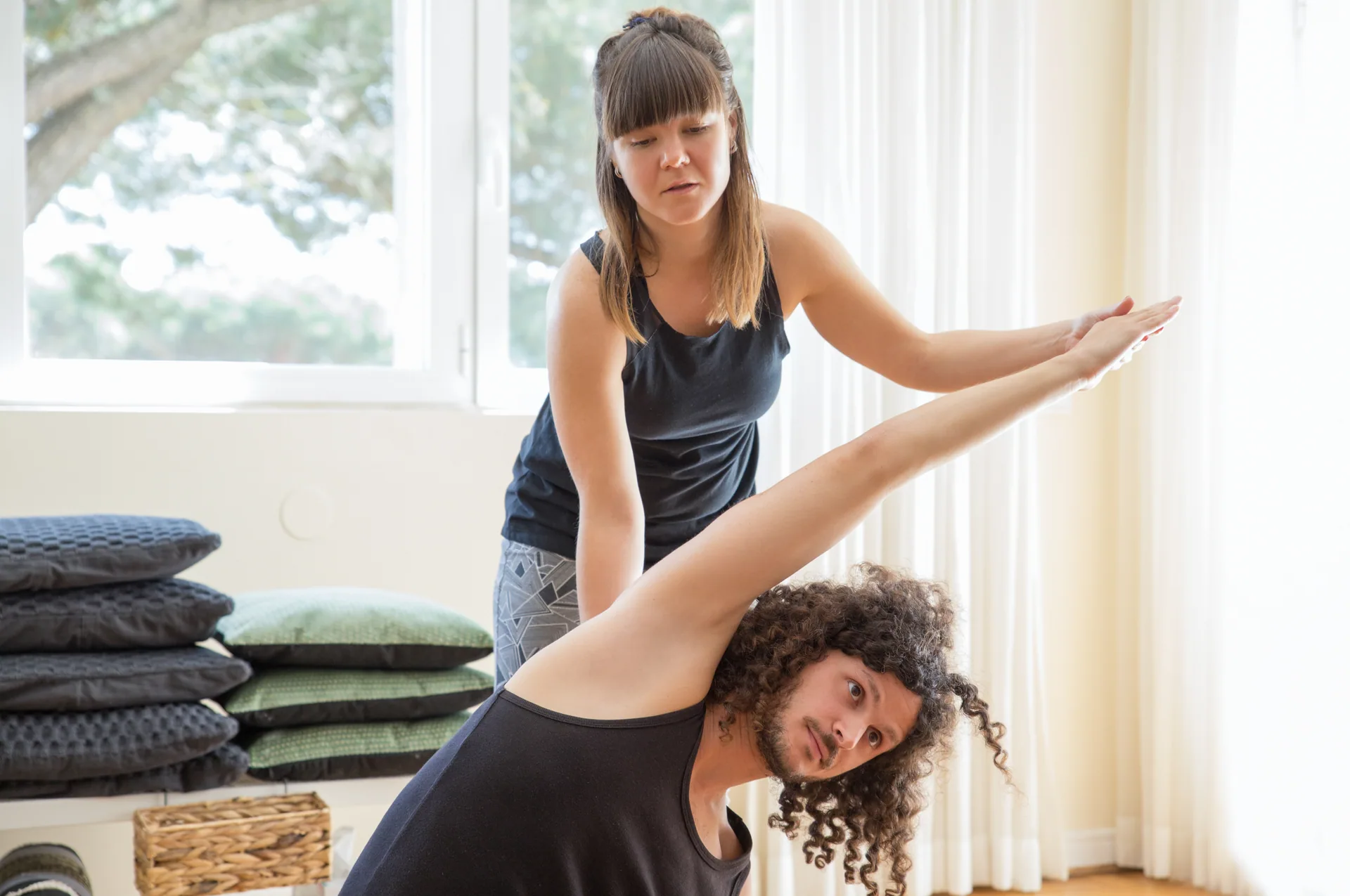 Instructor guiding a man with curly hair through a side stretch with raised arm in a bright studio with cushions nearby.