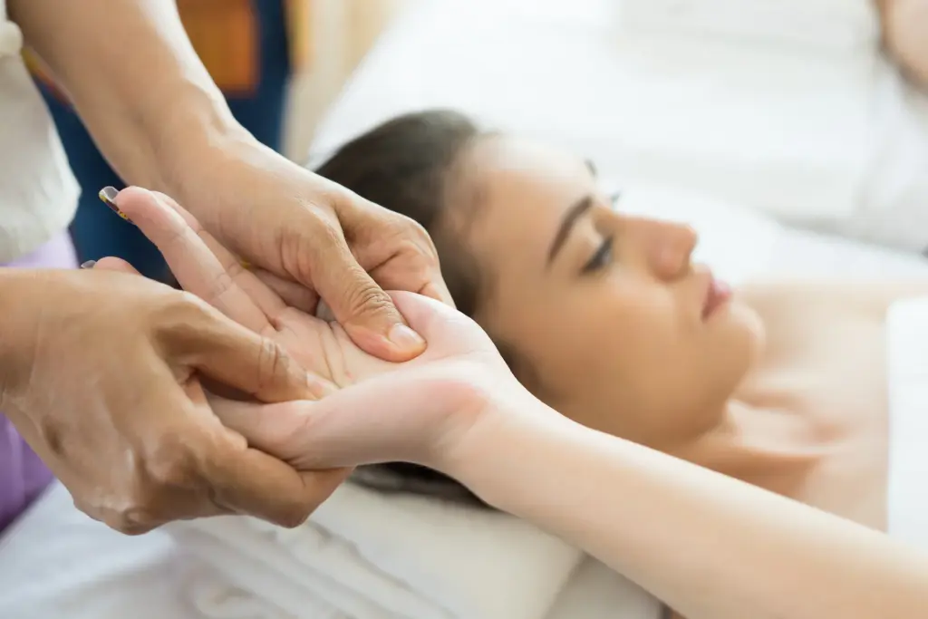 Massage therapist pressing thumbs into a client's palm while the client lies on a treatment table.