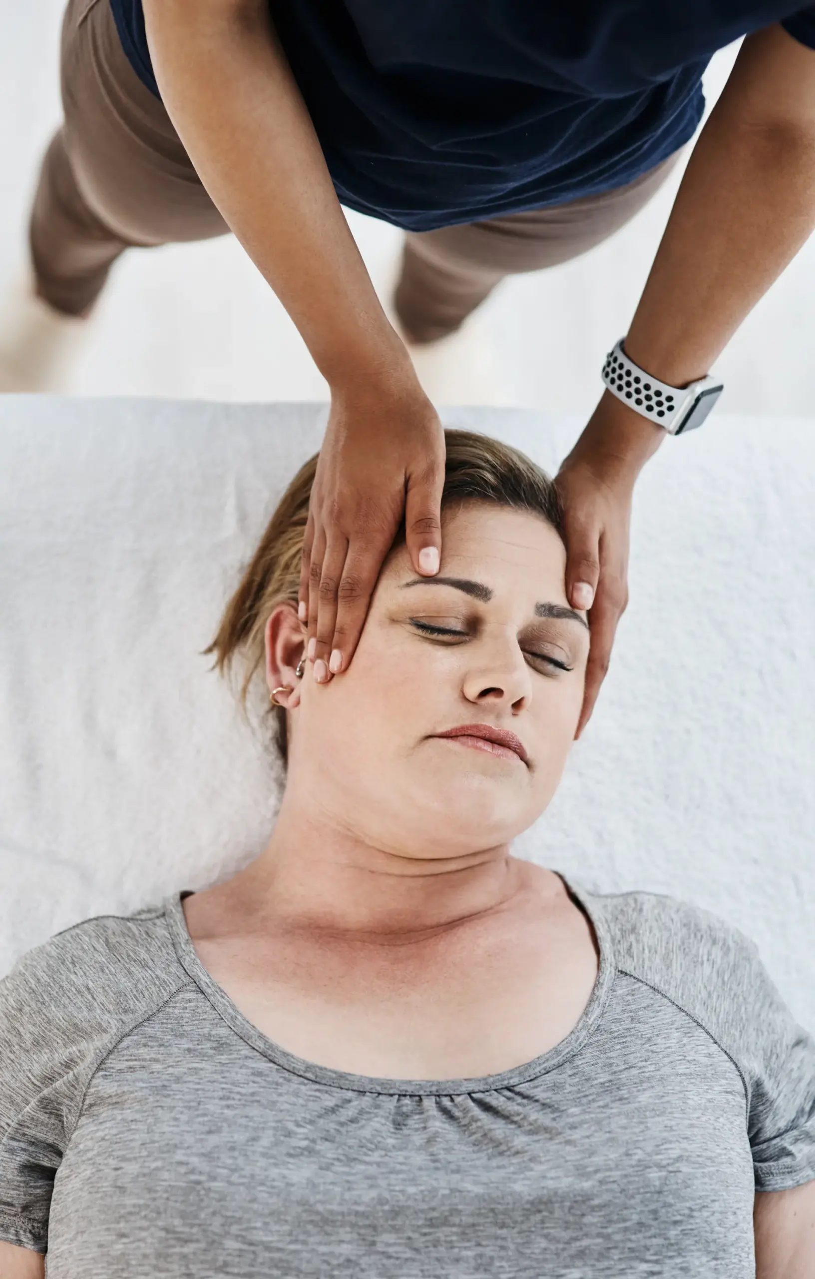 Overhead view of a therapist's hands resting on the temples of a woman lying on a massage table with eyes closed