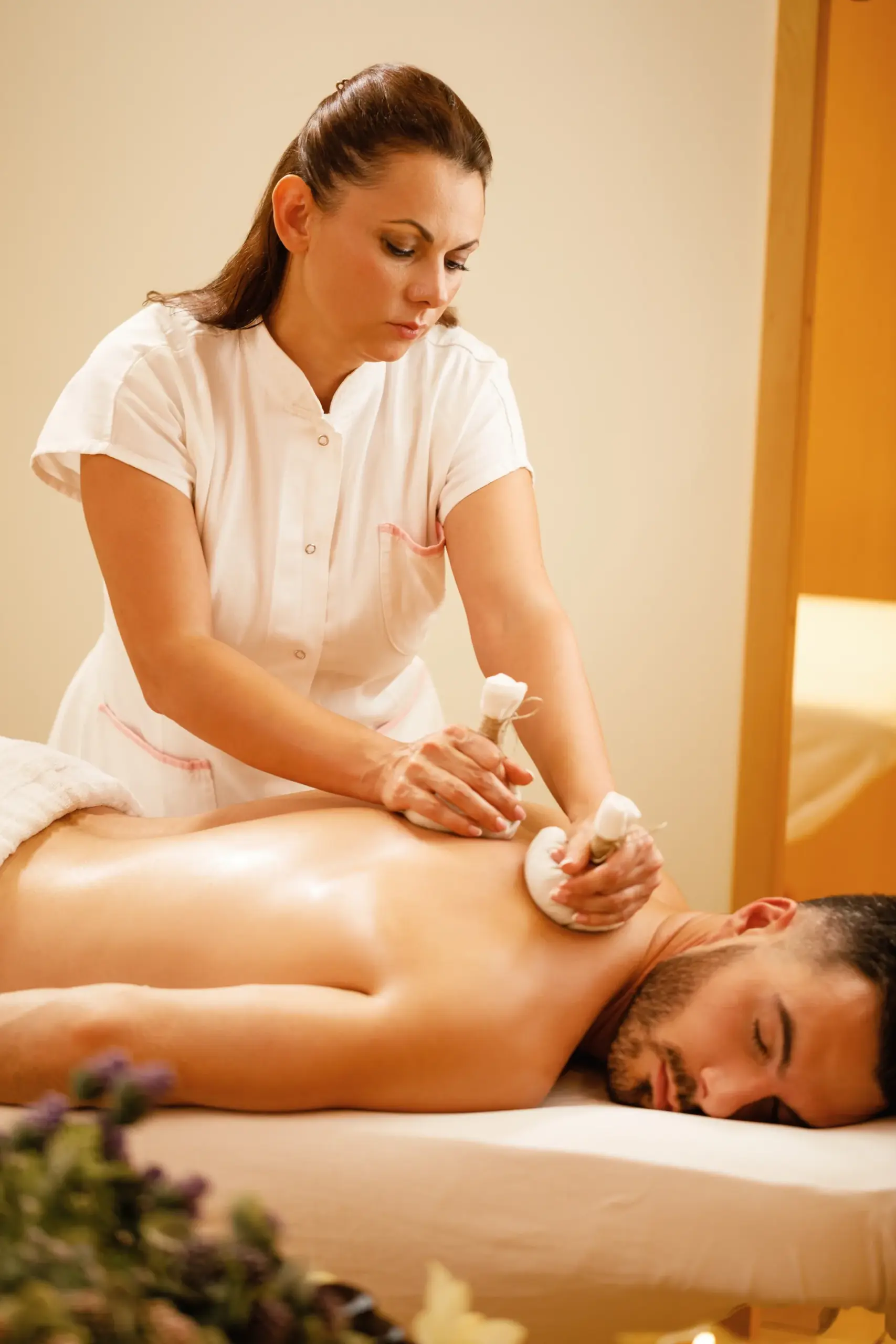 Massage therapist pressing two herbal compress balls into a man's back as he lies face down on a massage table.