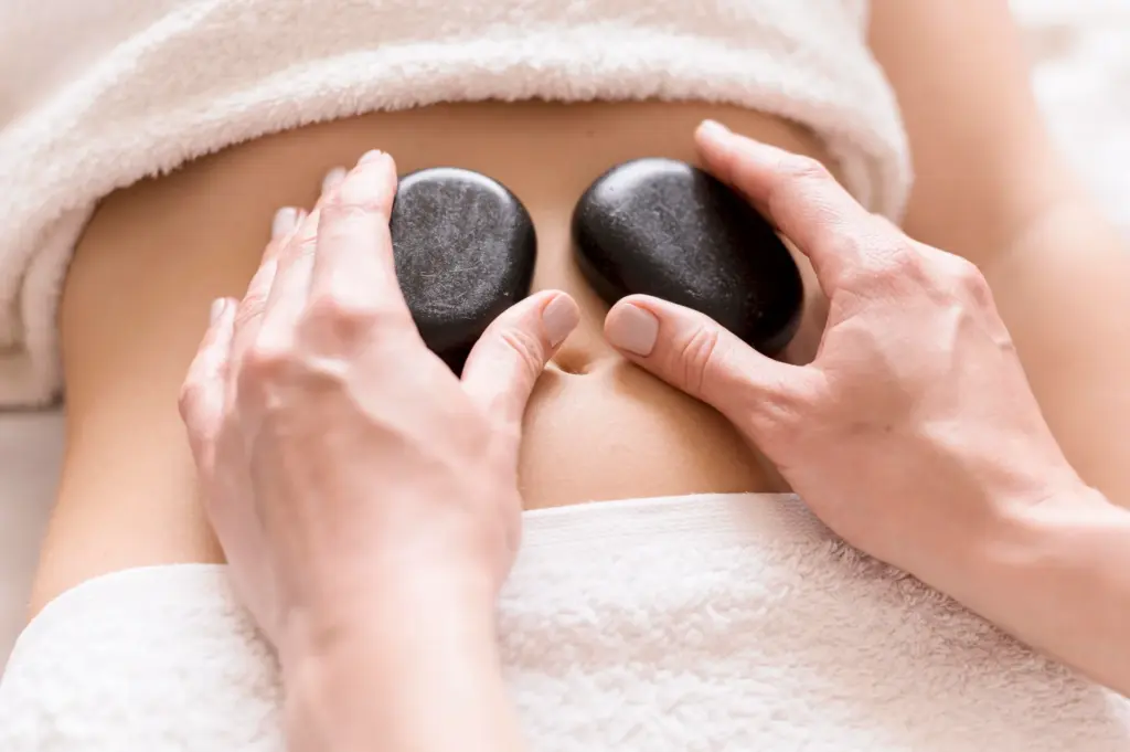 Massage therapist placing two smooth black basalt stones on a client's abdomen between white towels.