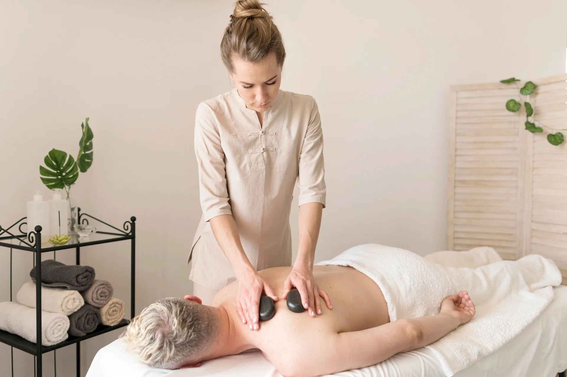 Massage therapist placing heated basalt stones on a client's upper back in a treatment room.