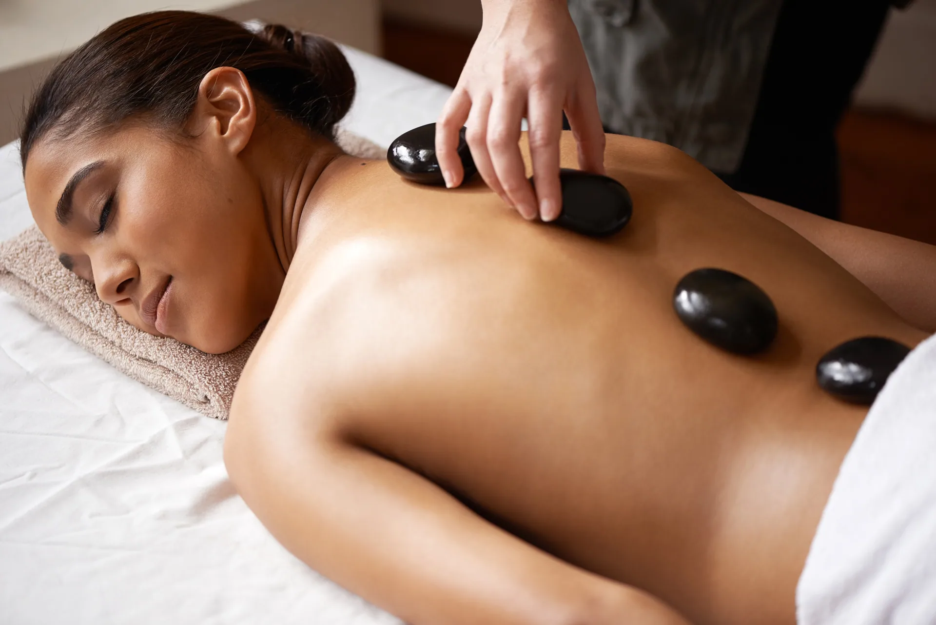 Woman lying face down on a massage table with heated basalt stones placed along her back by a therapist.