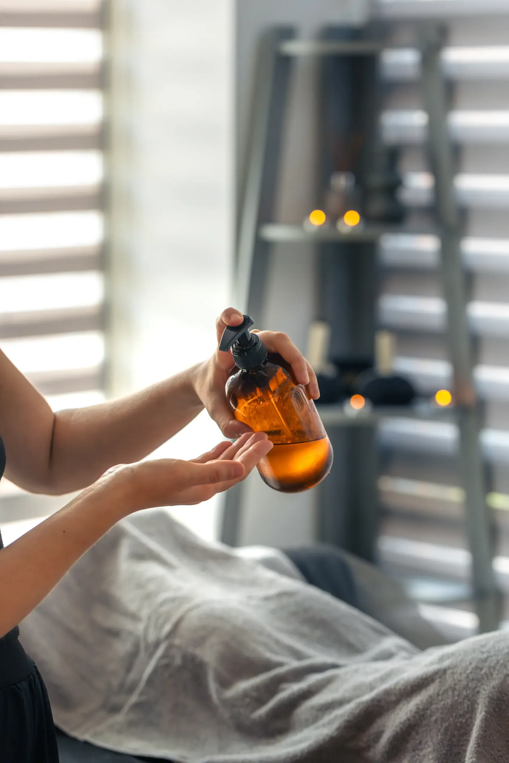 Therapist pressing an amber pump bottle to dispense massage oil into her hand beside a draped client on a massage table.
