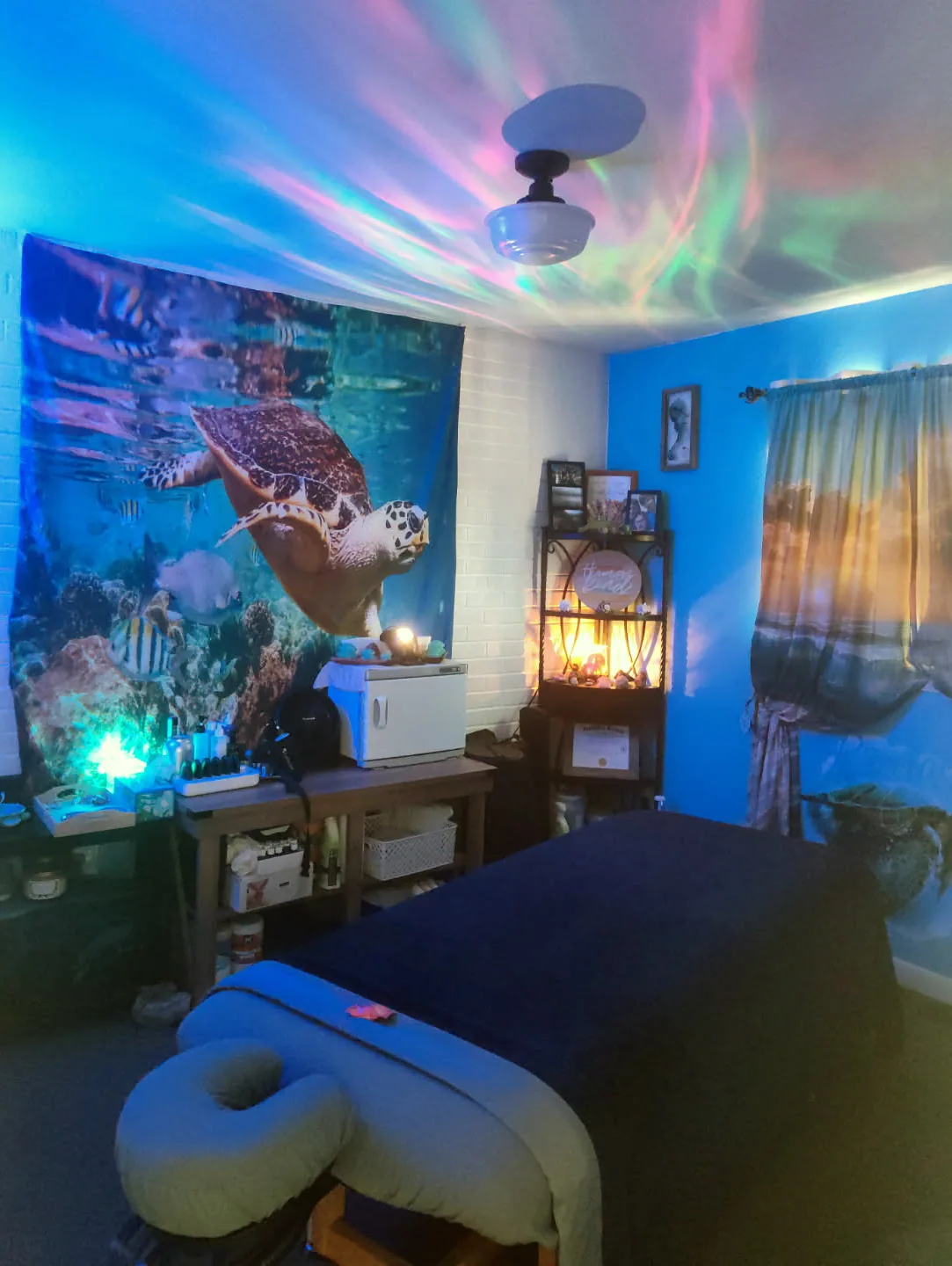 Massage room with a padded table, sea turtle tapestry, towel warmer, and colorful aurora lighting on the ceiling.