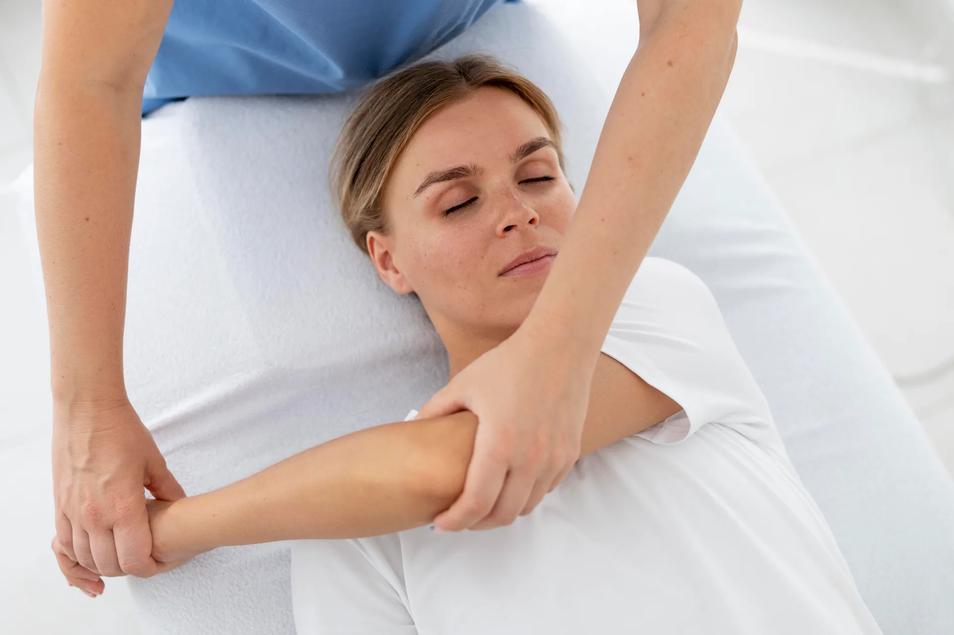 Therapist stretches a woman's arm across her body as she lies on a treatment table with eyes closed.