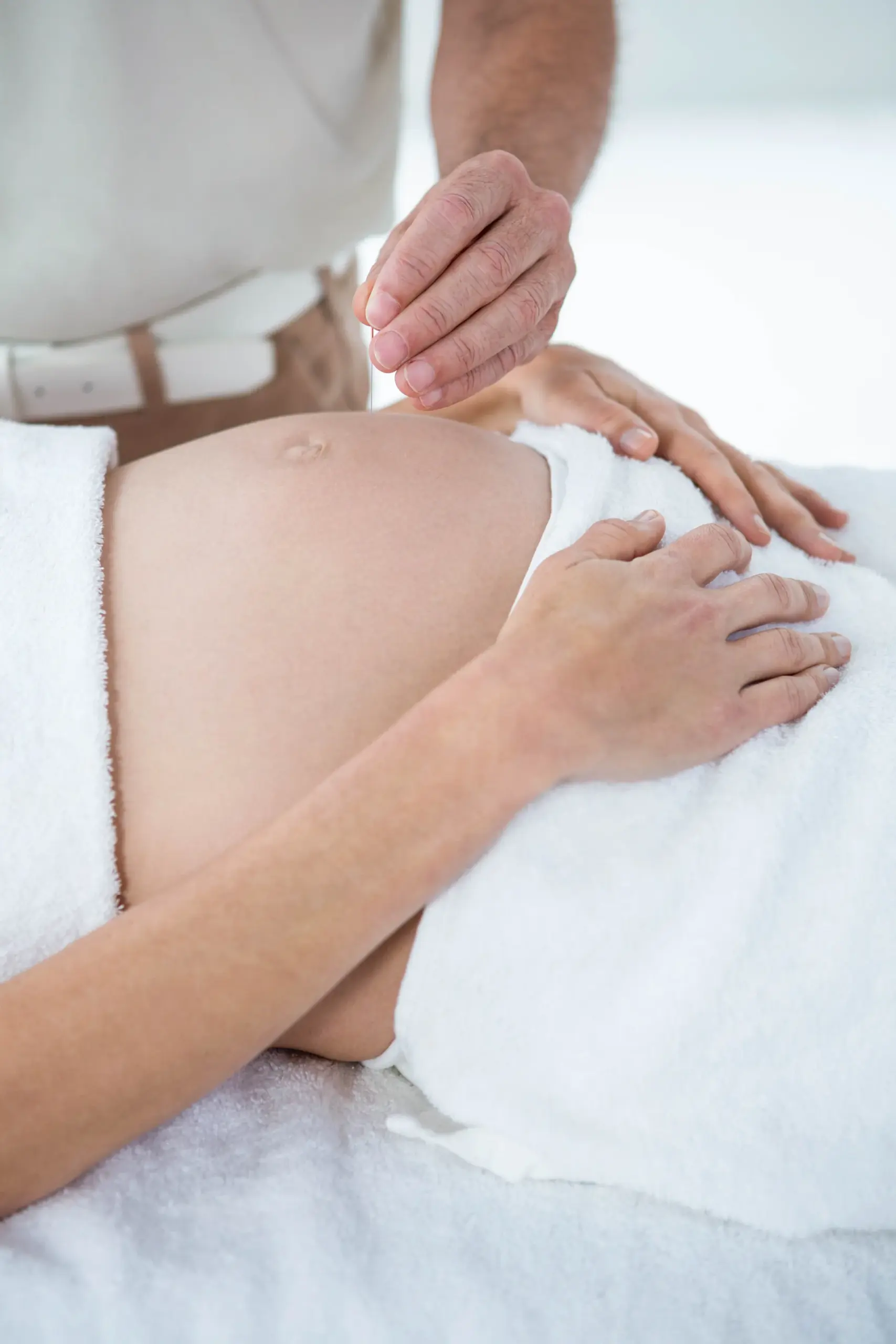 Practitioner holds an acupuncture needle above the bare belly of a pregnant woman lying on a treatment table.