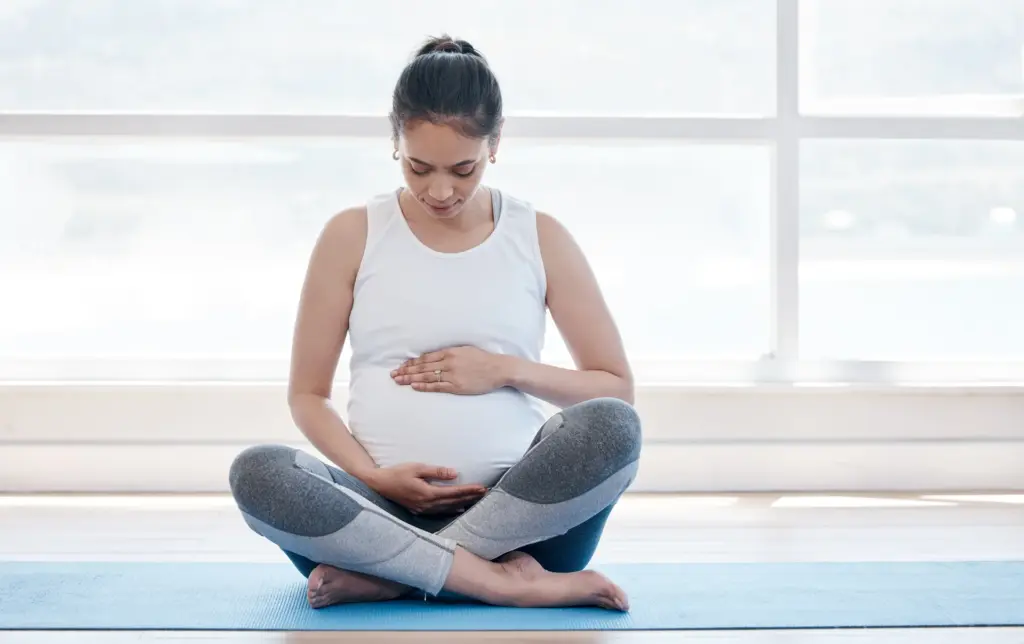Pregnant woman sitting cross-legged on a blue yoga mat, hands resting on her belly, in front of a bright window.