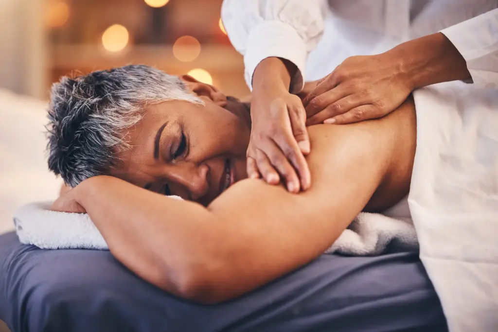 Woman with short gray hair lying on a massage table while a therapist massages her shoulder.