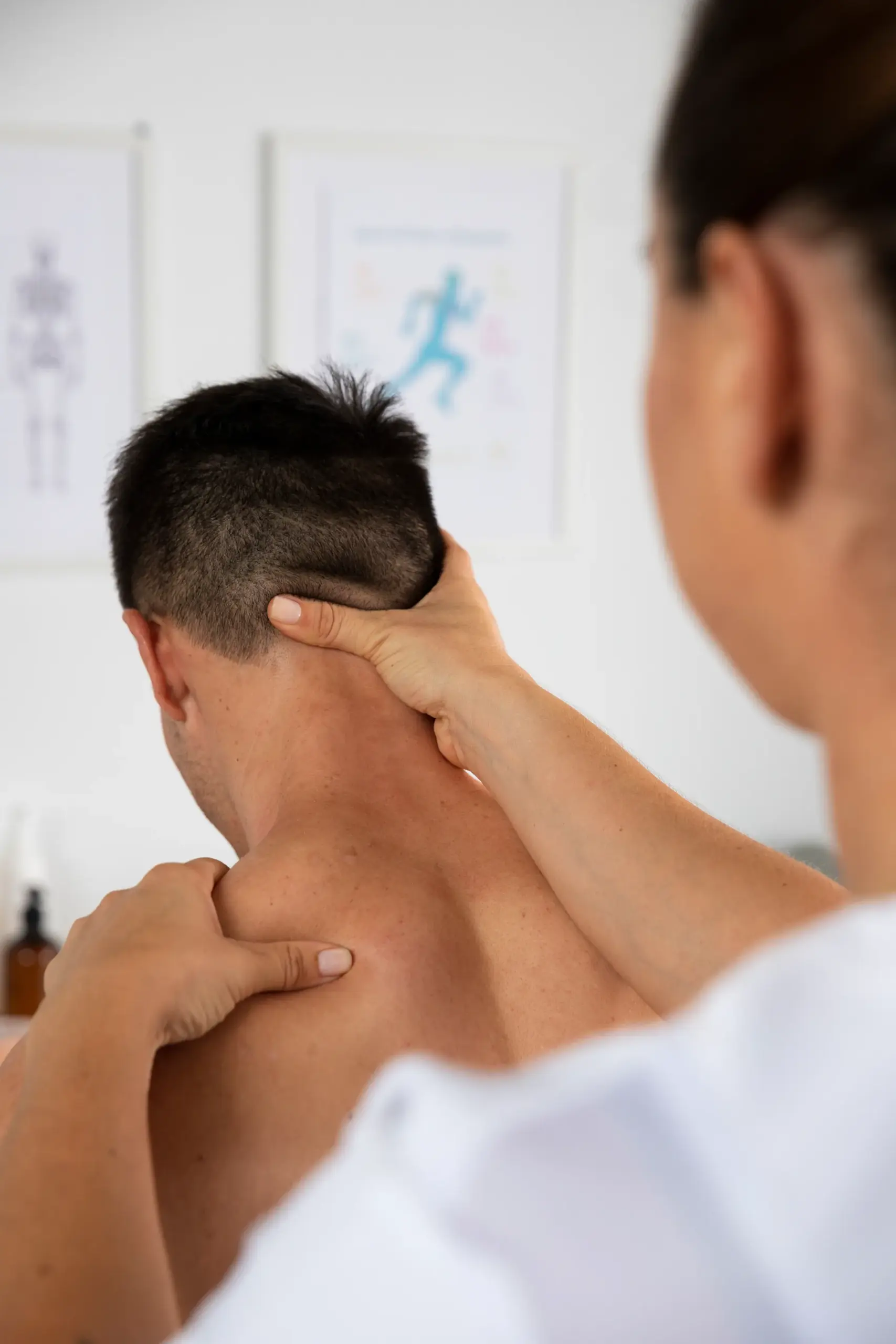 Massage therapist standing behind a seated male client, working on his neck and shoulder in a treatment room.