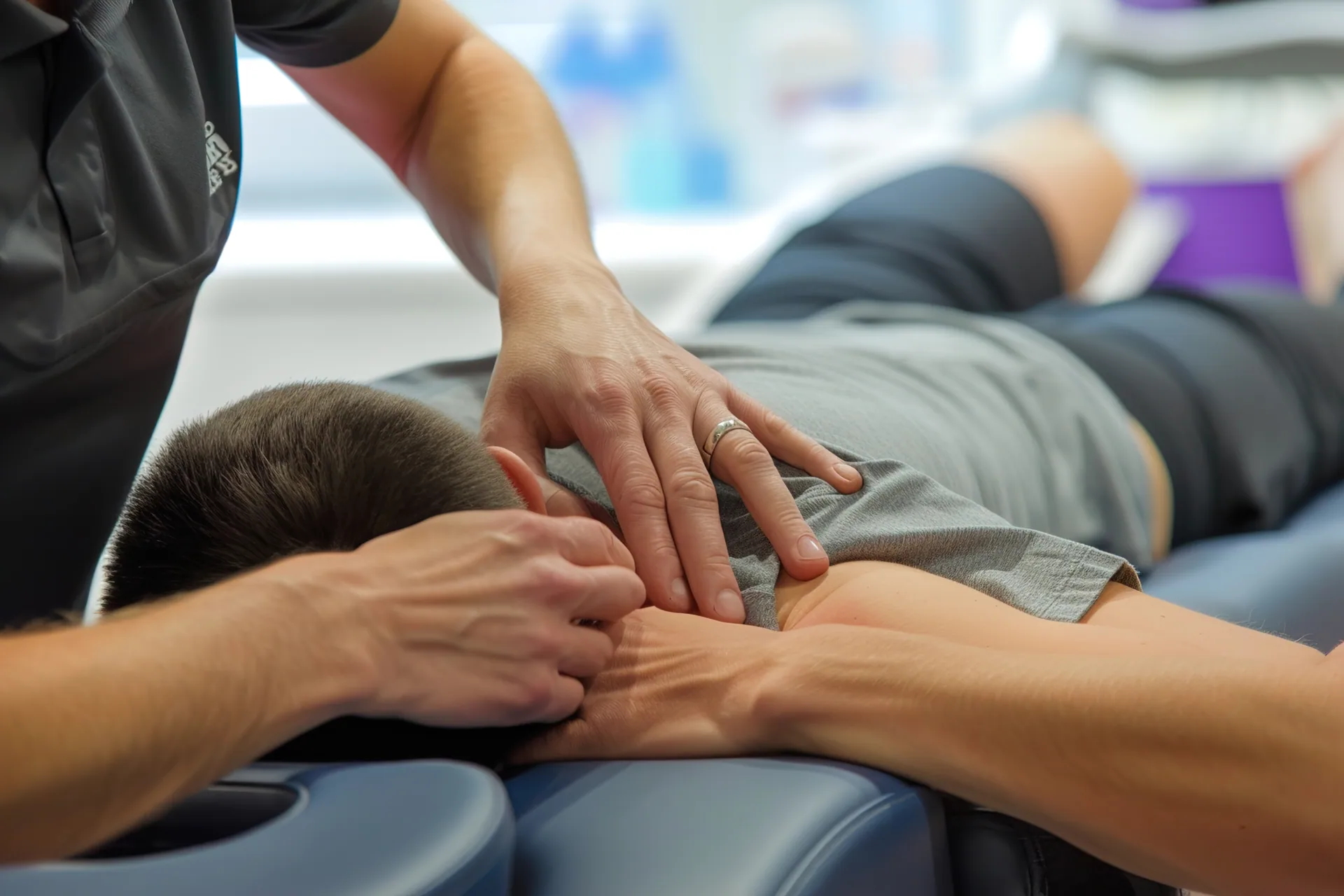 Massage therapist using both hands to work on a clothed client's shoulder and neck while the client lies face down on a