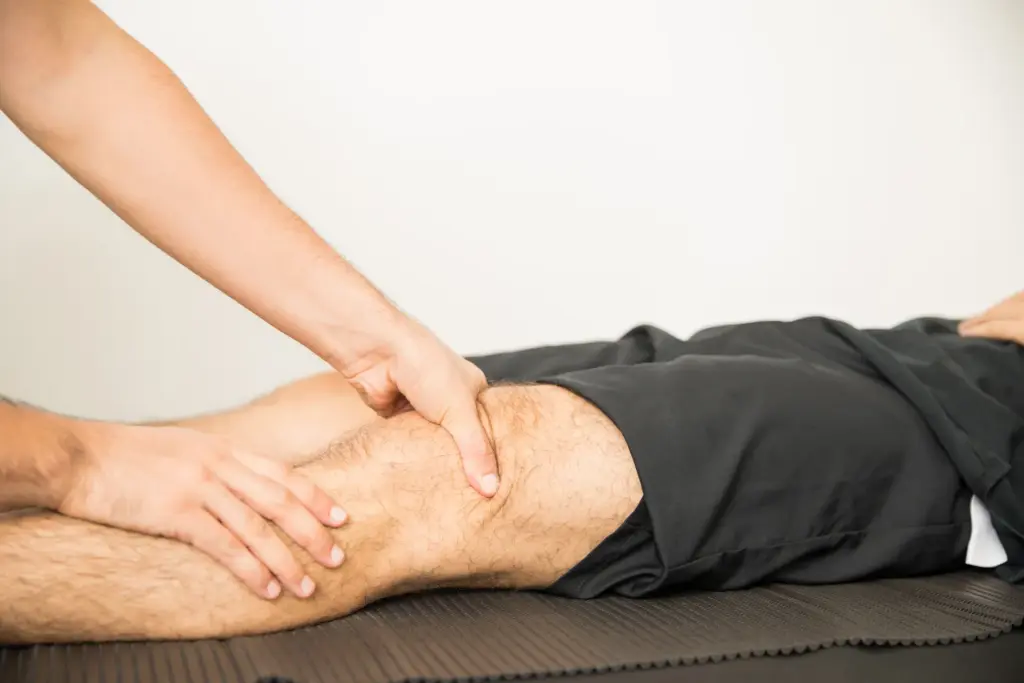 Massage therapist using both hands to work on a male client's knee and thigh while he lies on a treatment table.