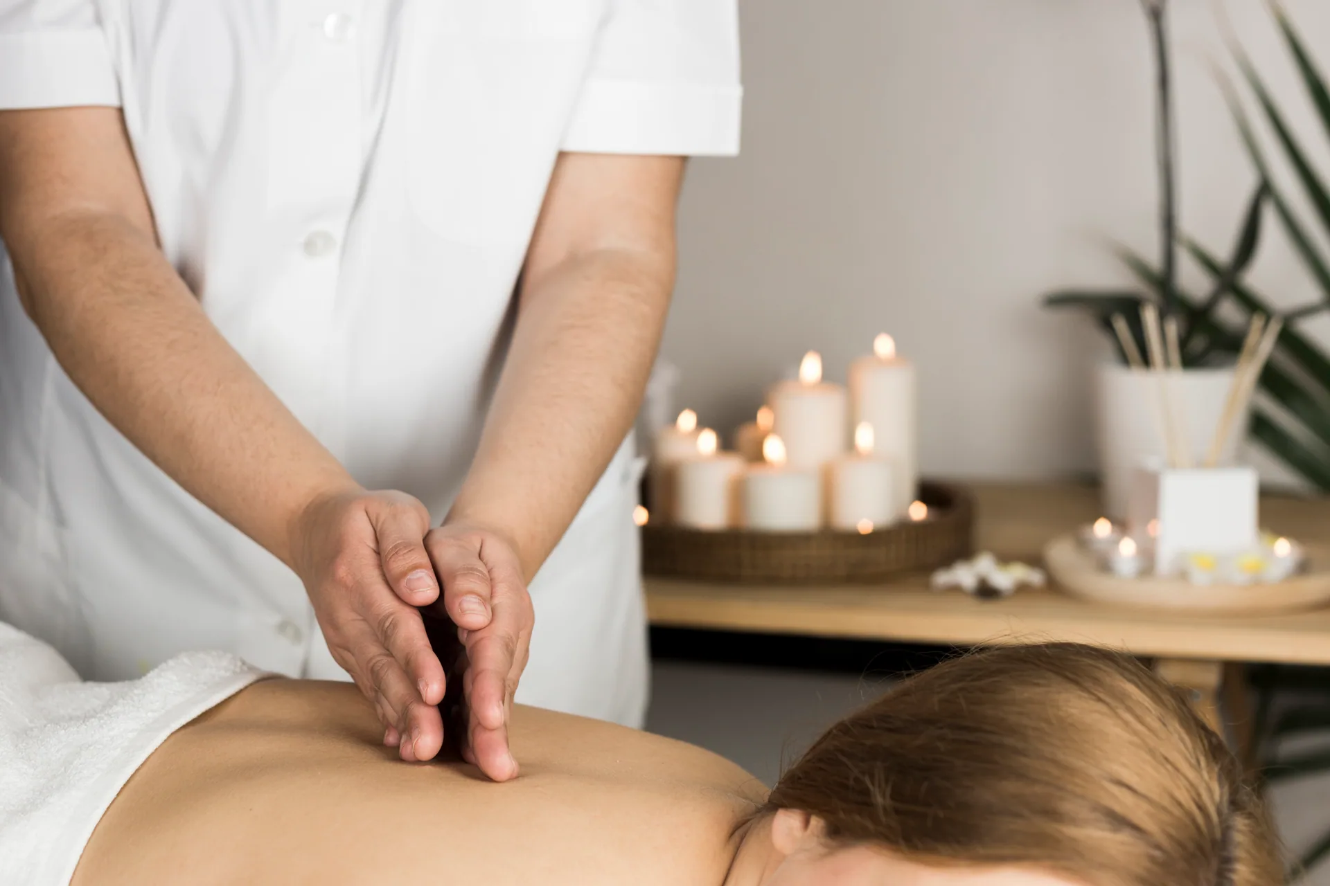 Massage therapist in white uniform performing a back massage on a woman lying on a table, with lit candles in the background.