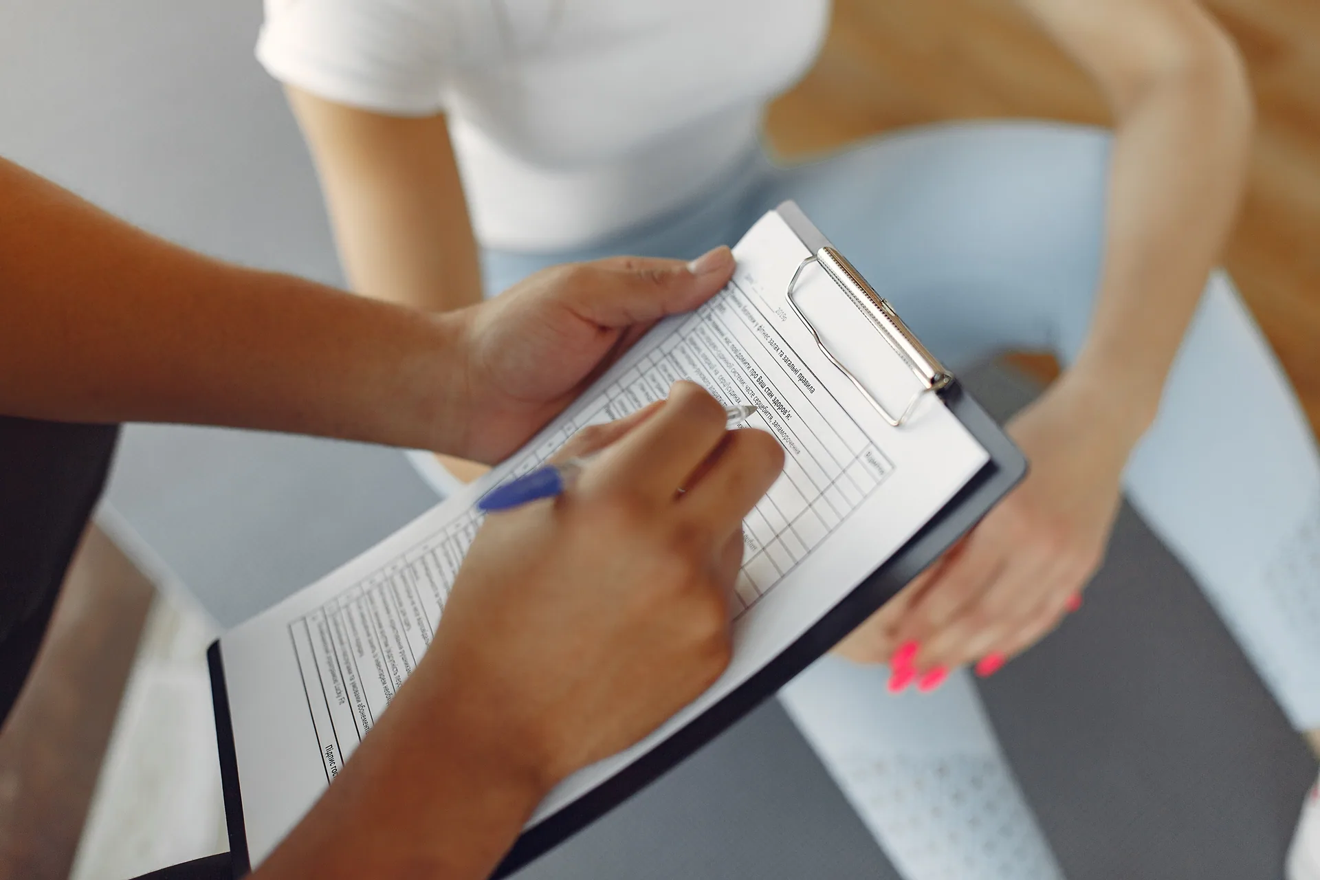 Trainer holding a clipboard and pen fills out an assessment form while a client sits on an exercise mat.